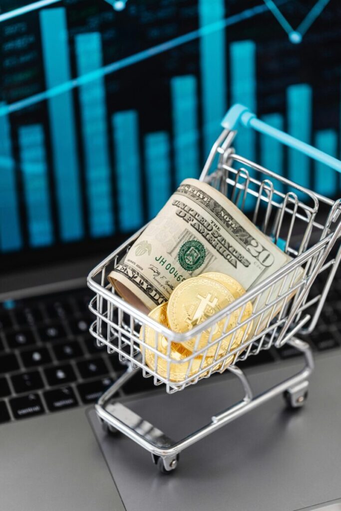 Shopping cart with bitcoin and cash on laptop with financial graph backdrop.