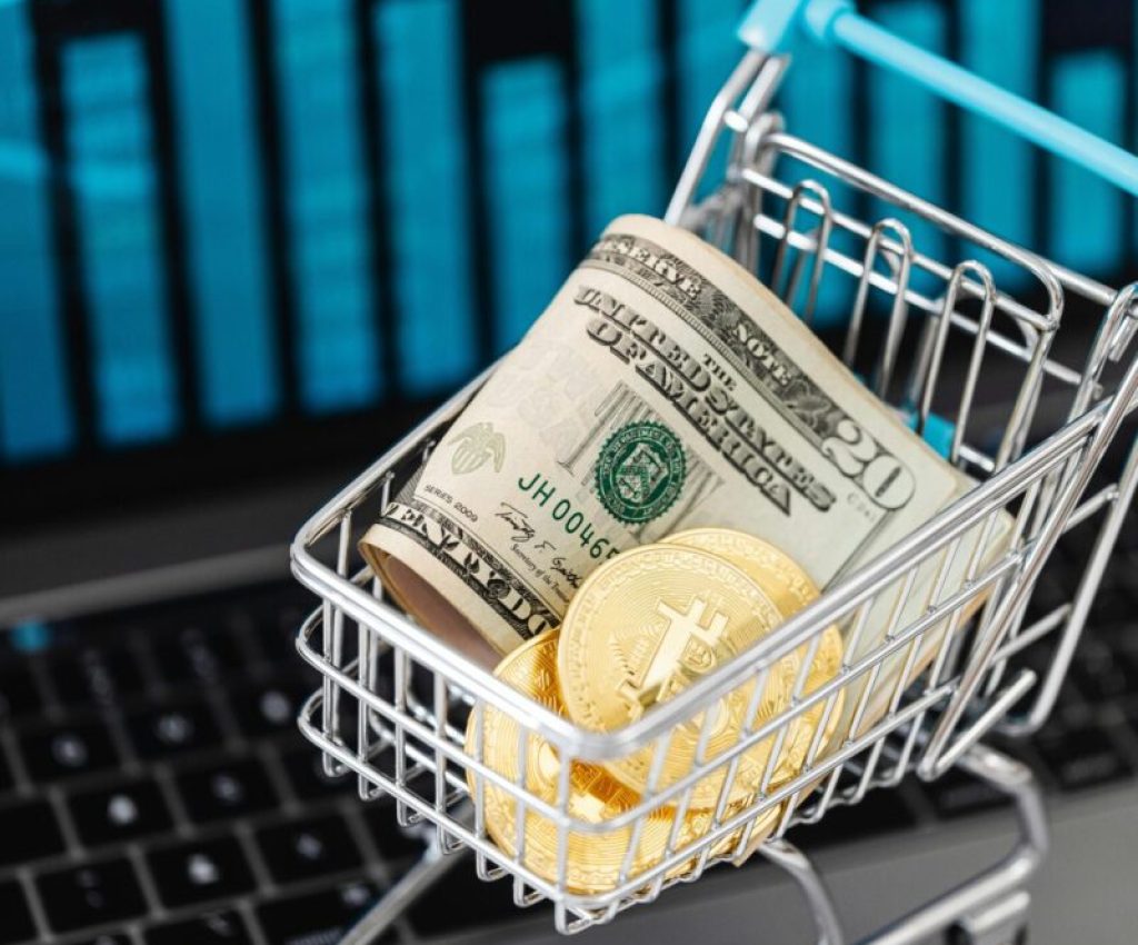 Shopping cart with bitcoin and cash on laptop with financial graph backdrop.